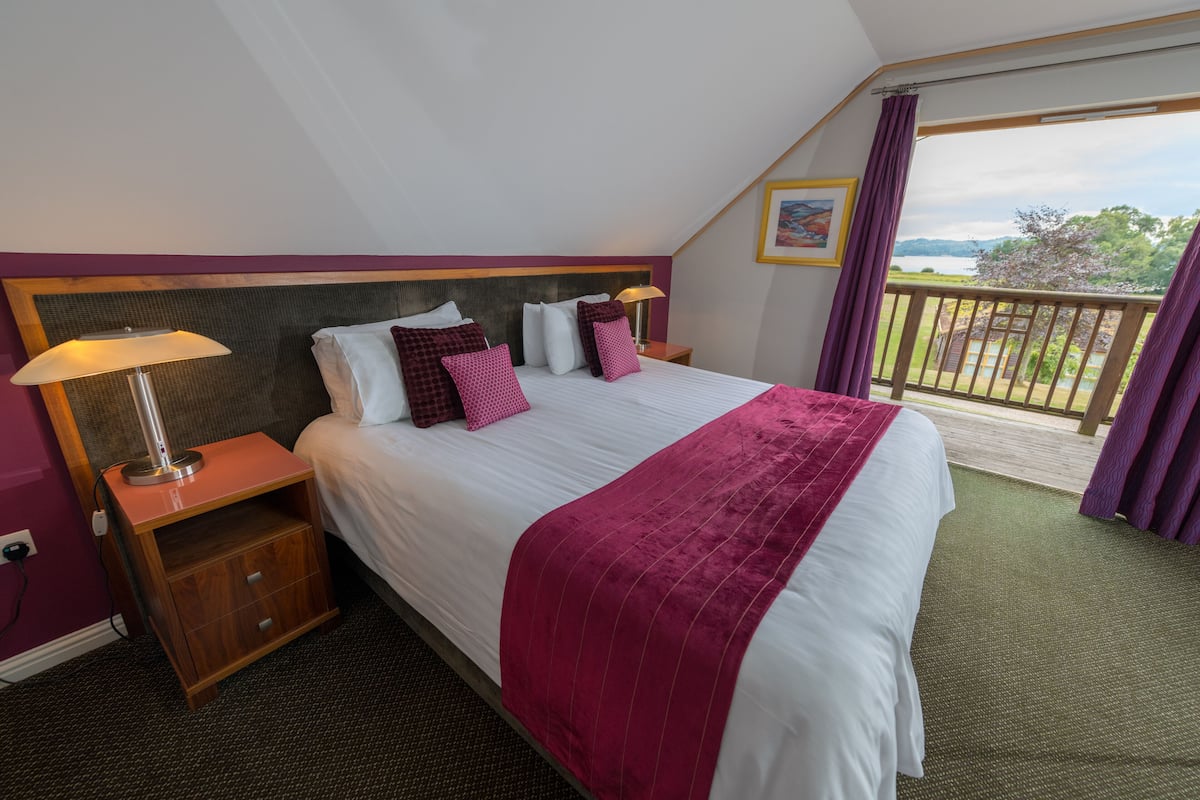 A hotel room with a large bed, a nightstand, a lamp, and a view of a balcony with a railing.