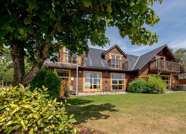 A two-story wooden house with a balcony, large windows, and a well-maintained lawn. - Loch Lomond Waterfront Luxury Lodges (Balmaha)