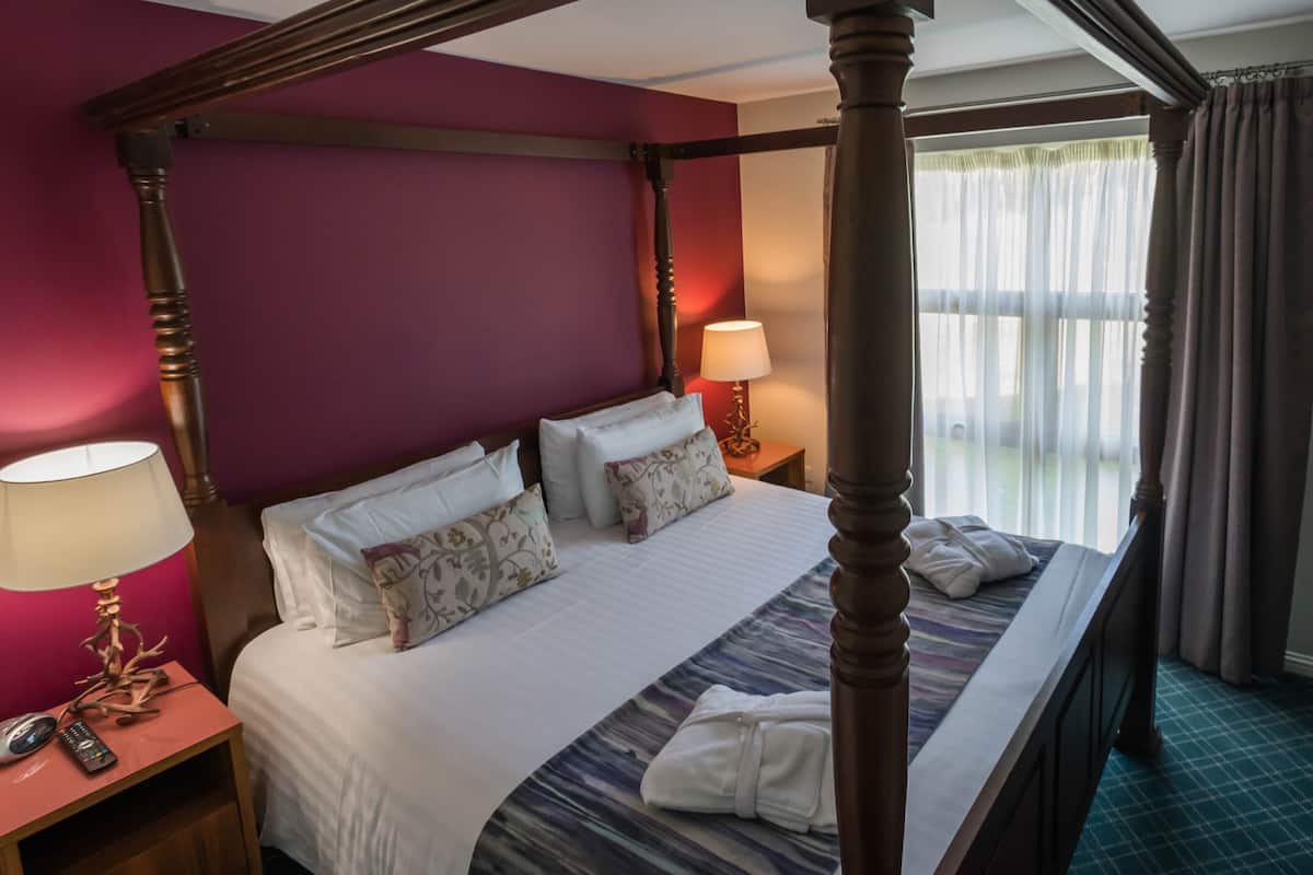 A four-poster bed with white linens, a bedside table with a lamp, and a view of the outdoors through sheer curtains.