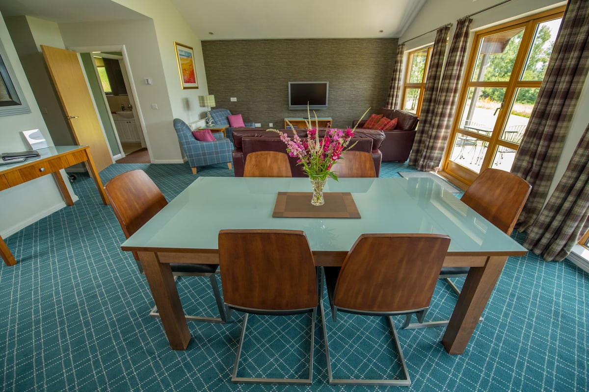 A dining area with a glass table and wooden chairs, a living room with a sofa and a TV, and a kitchen in the background.