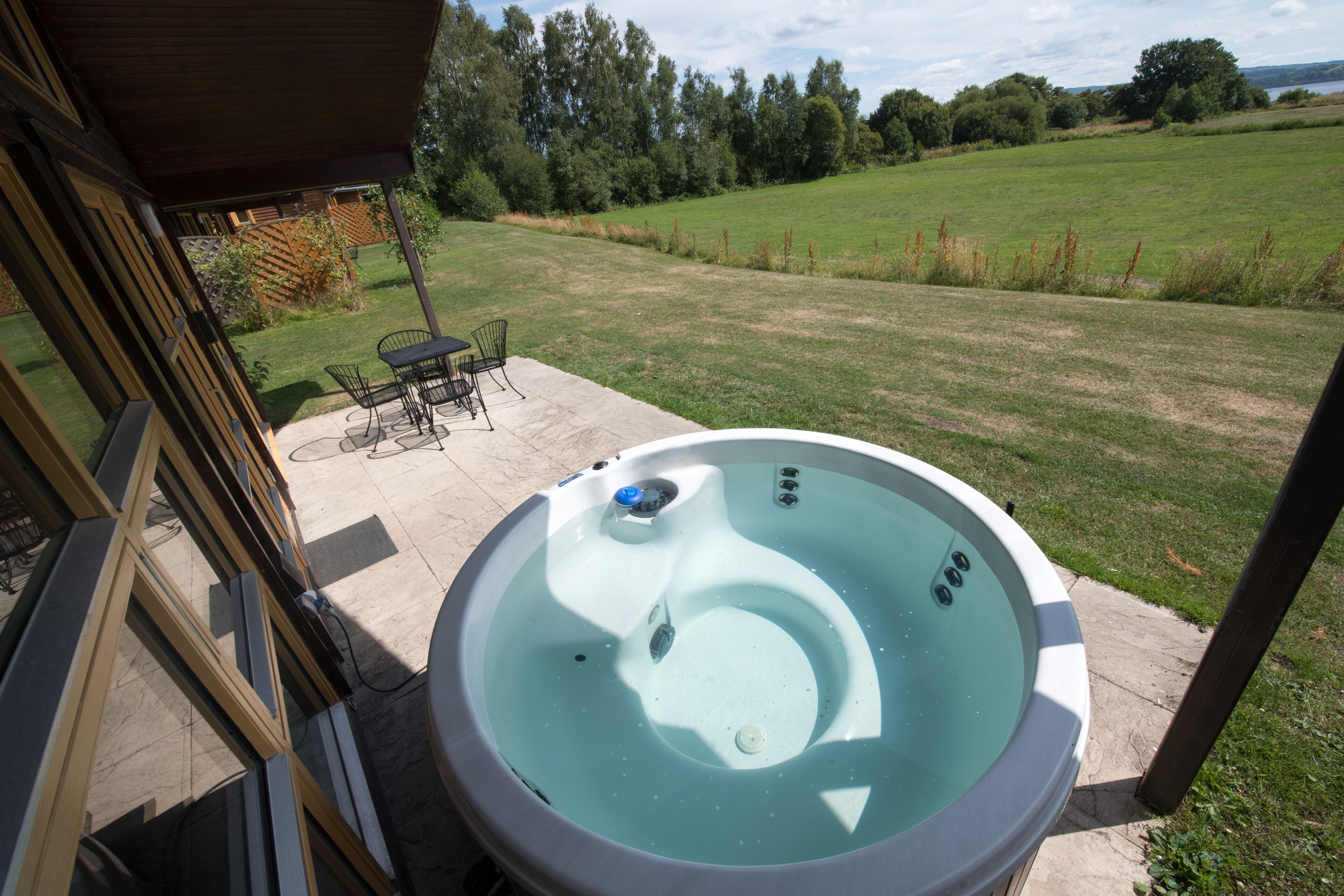 A hot tub in a backyard with a patio table and chairs, surrounded by a grassy lawn and trees.