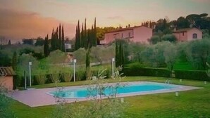 A swimming pool surrounded by a grassy area with a view of distant buildings and trees under a clear sky. - Gli Archi (Fauglia)