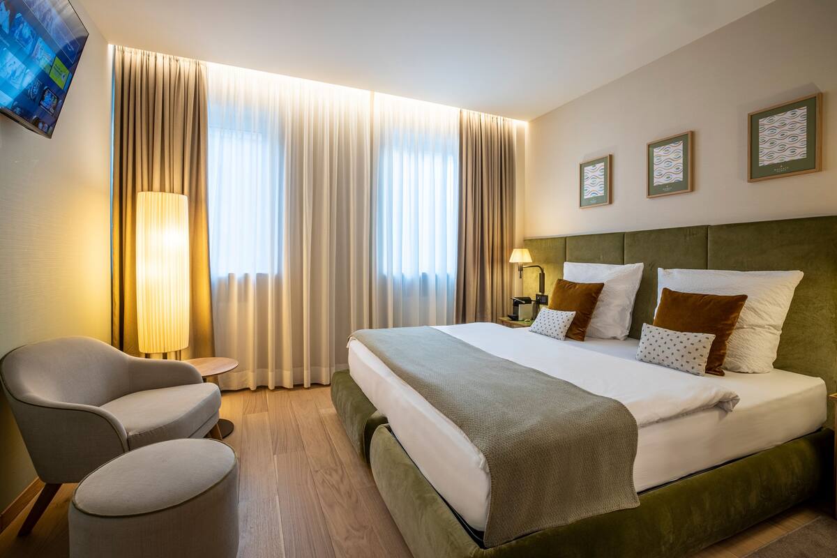 Hotellihuone, jossa on suuri sänky, tuoli, pieni pöytä ja seinälle asennettu televisio.