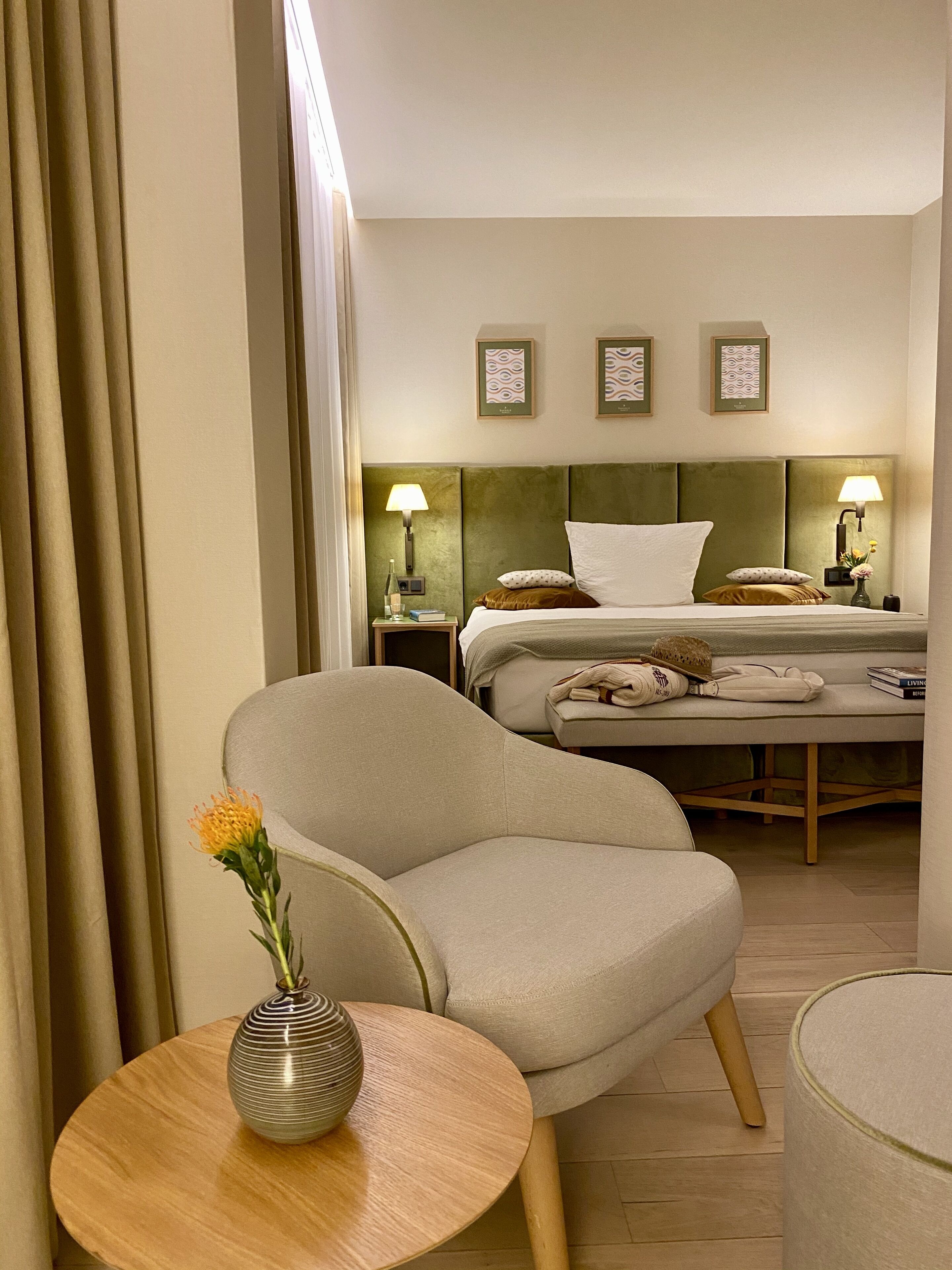 Habitación de hotel con cama, una silla, una mesita con un jarrón y una mesita de noche con lámpara.