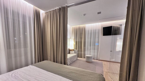 Una habitación de hotel moderna con una cama grande, una zona de estar con sofá y sillón, una mesita redonda y una televisión.