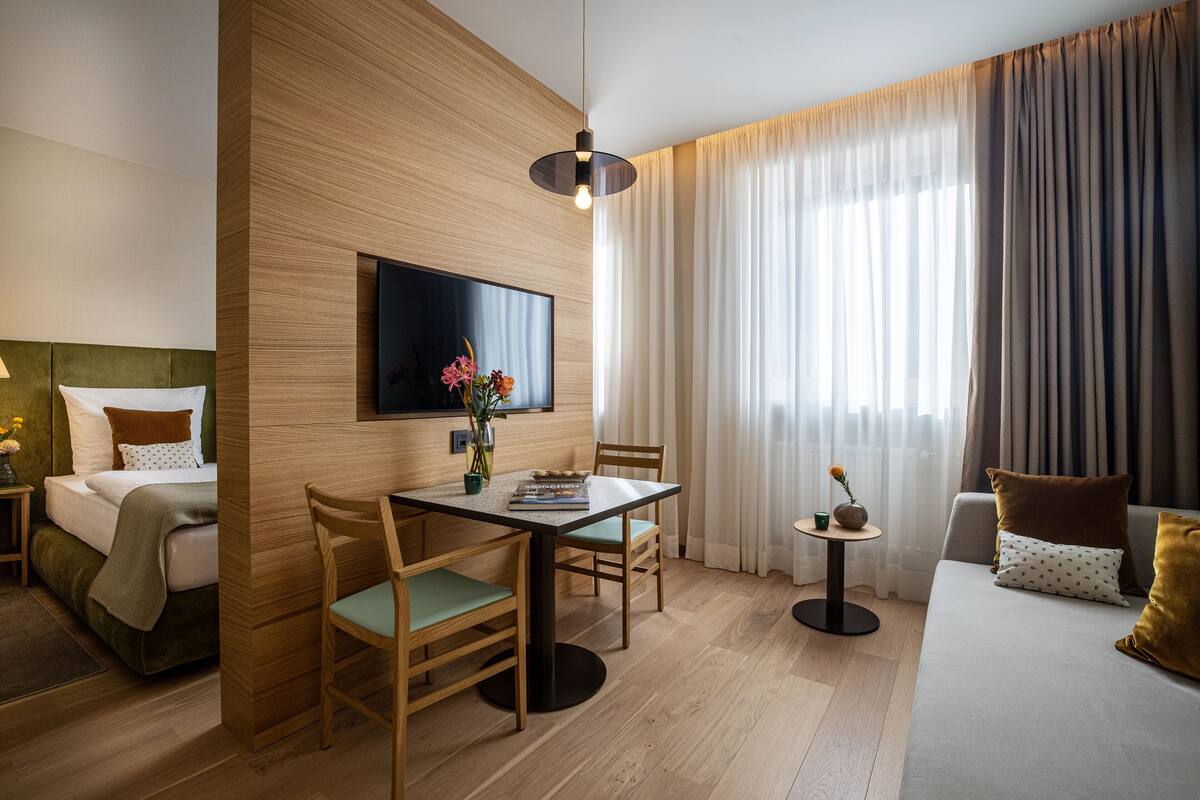 Hotellihuone, jossa on sänky, ruokapöytä tuoleineen, seinälle asennettu televisio, pieni sivupöytä maljakko täynnä kukkia ja sohva koristetyynyineen.