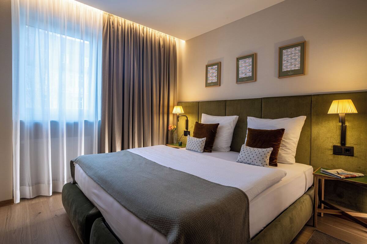 Hotellihuone, jossa on suuri sänky, yöpöytä lampun kanssa ja ikkuna, jossa on verhot.