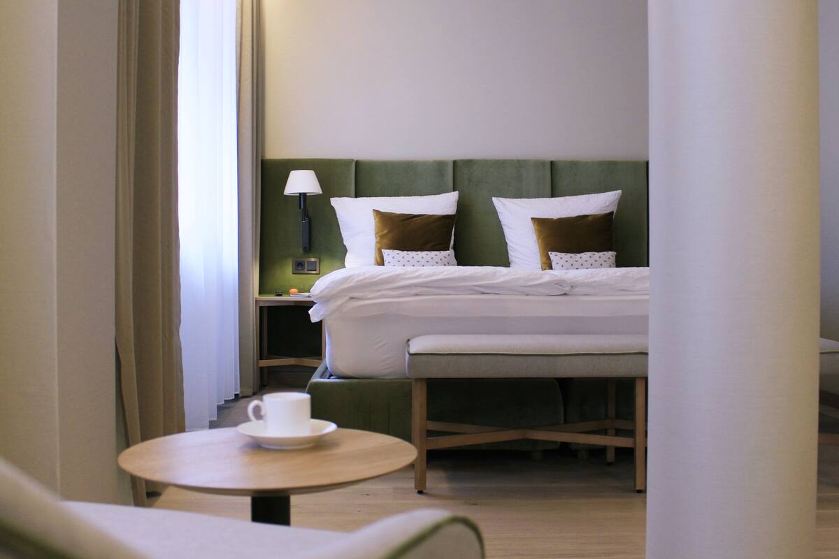Hotellihuoneessa on sänky, yöpöytä lampun kanssa, pieni pyöreä pöytä, jossa on kuppi, ja tuoli.