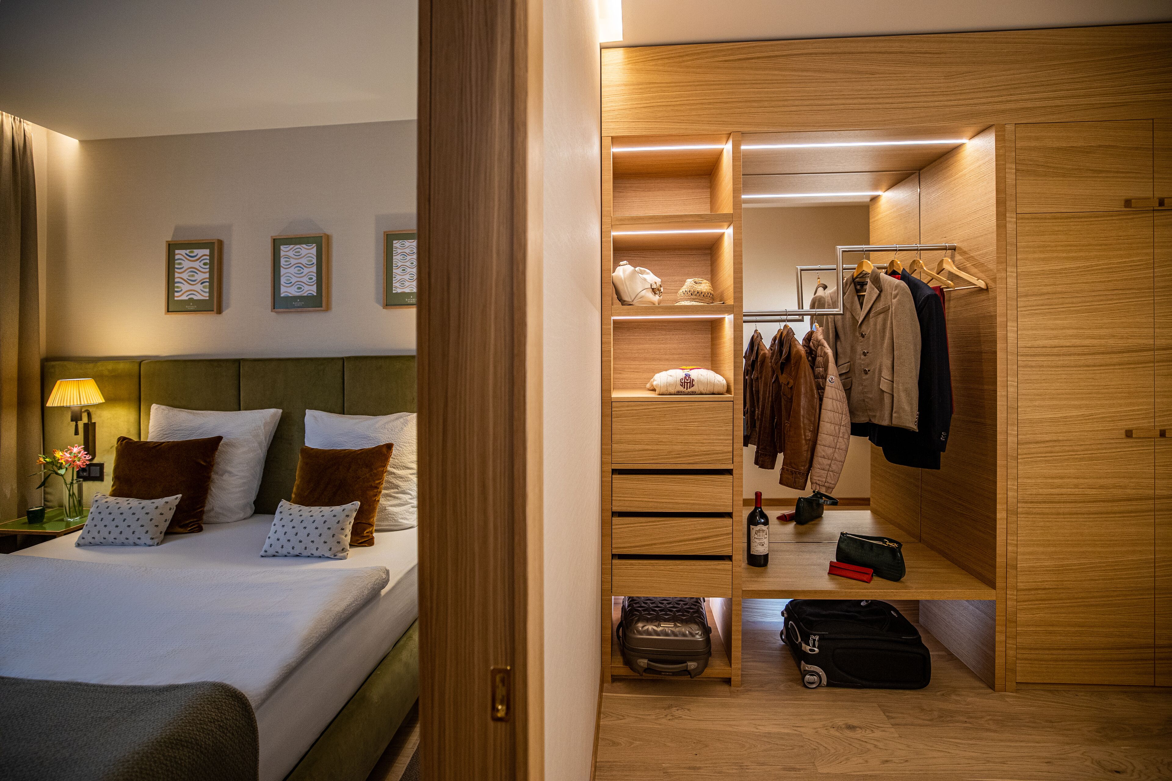 Habitación de hotel con una cama bien hecha, un armario de madera con ropa colgada y una mesita con una botella y una bolsa.