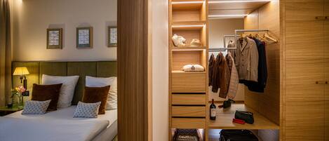 Habitación de hotel con una cama bien hecha, un armario de madera con ropa colgada y una mesita con una botella y una bolsa.