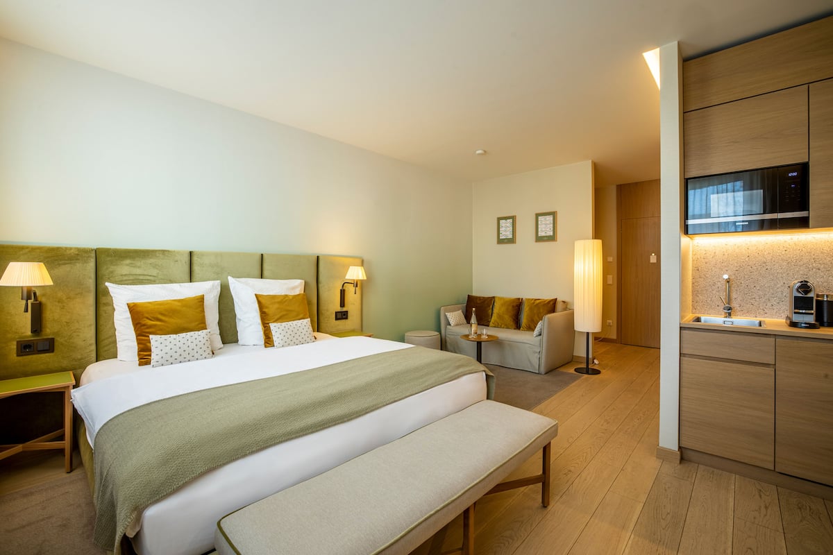 Hotellihuone, jossa on sänky, yöpöydät, penkki, sohva, pieni pöytä ja pieni keittotila.