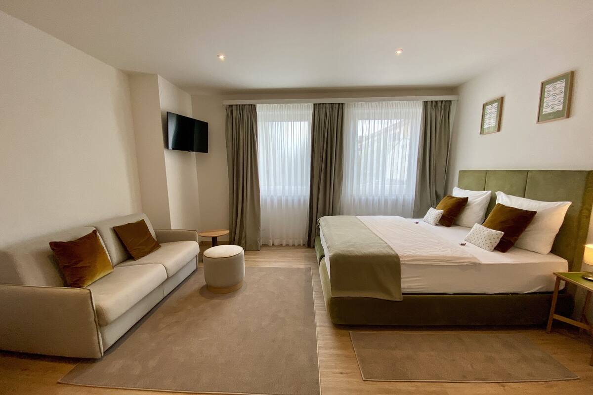 Hotellihuone, jossa on sänky, sohva, televisio ja ikkuna, jossa on verhot.