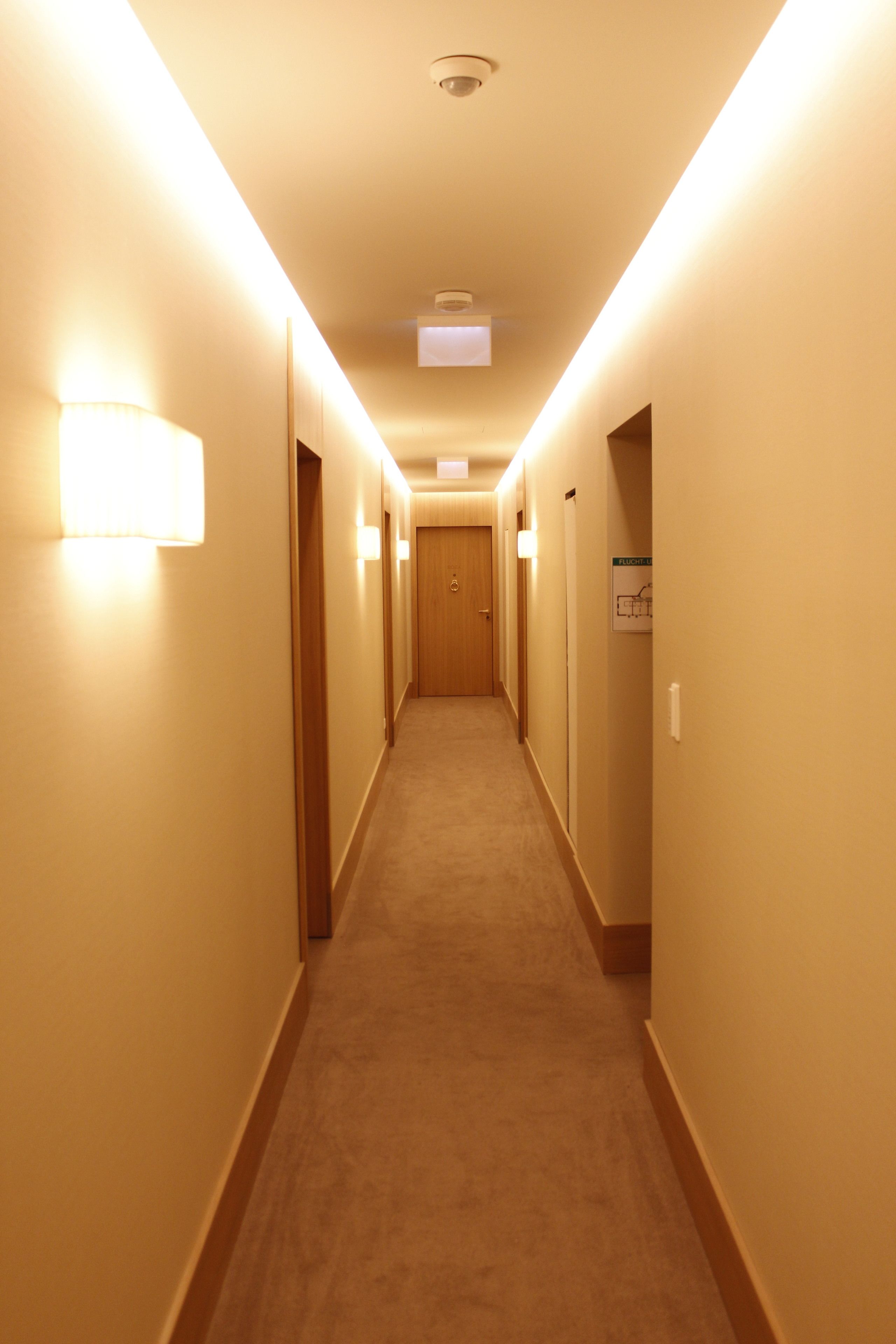 Un couloir d'hôtel avec des revêtements de sol en moquette, des portes en bois et des luminaires fixés au mur.