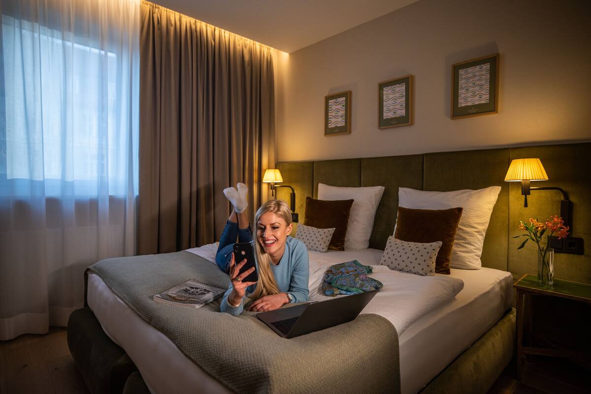 Hotellihuone, jossa on sänky, kannettava tietokone, älypuhelin sekä yöpöytä, jossa on lamppu ja maljakko täynnä kukkia.