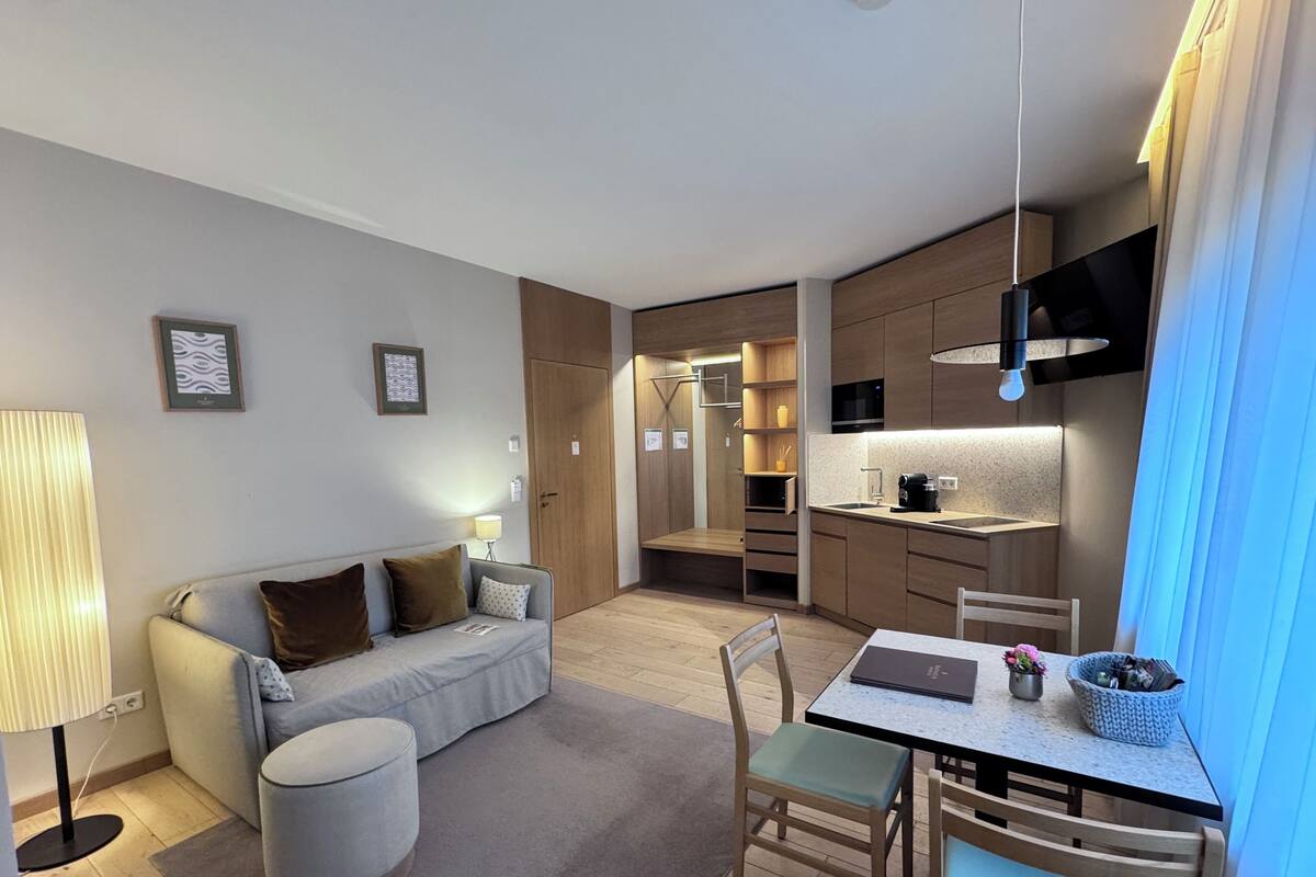 Moderni hotellihuone, jossa on sohva, ruokapöytä tuoleineen, pieni keittiö ja erillinen vaatehuone.