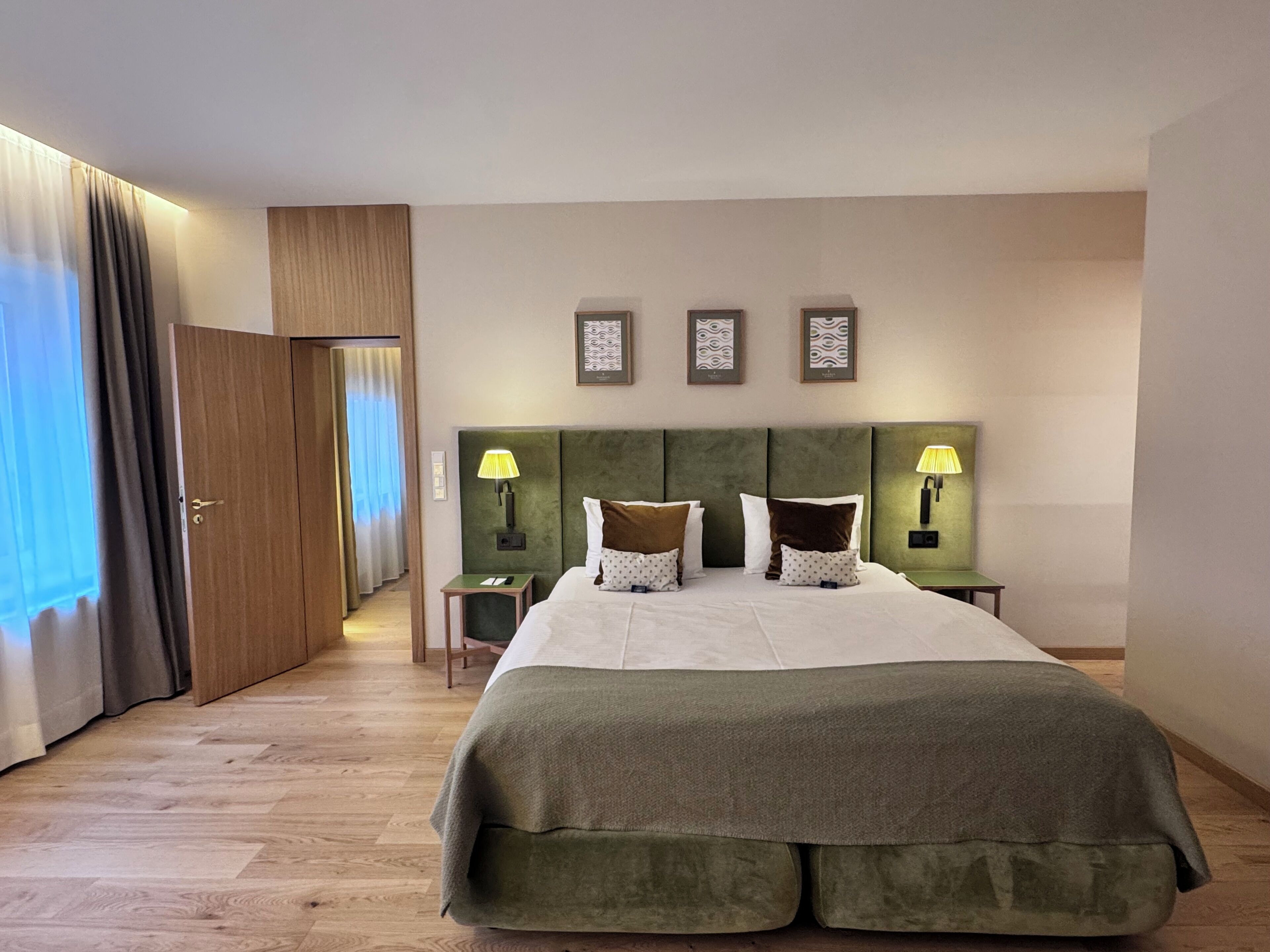 A hotel room with a large bed, two bedside tables with lamps, a door leading to another room, and a view of the outside.