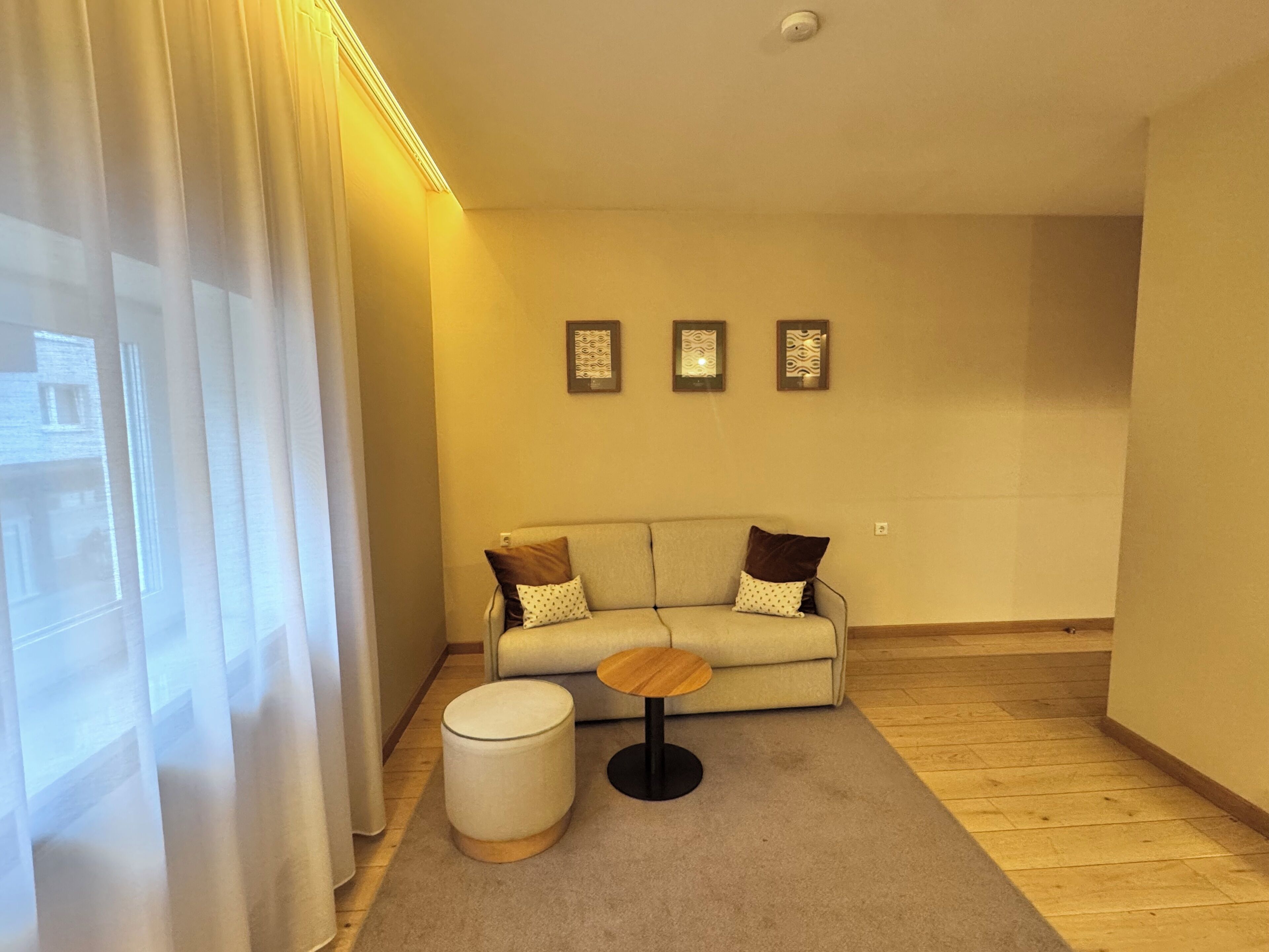 Ein modernes Wohnzimmer mit einer Couch, einem runden Couchtisch und einem kleinen Hocker. An der Wand hängen drei gerahmte Bilder.