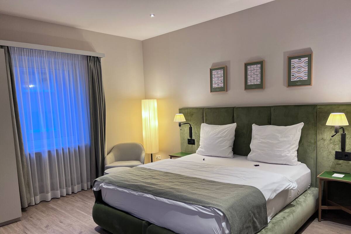 Hotellihuone, jossa on suuri sänky, kaksi yöpöytävalaisinta, tuoli ja ikkuna, jossa on verhot.