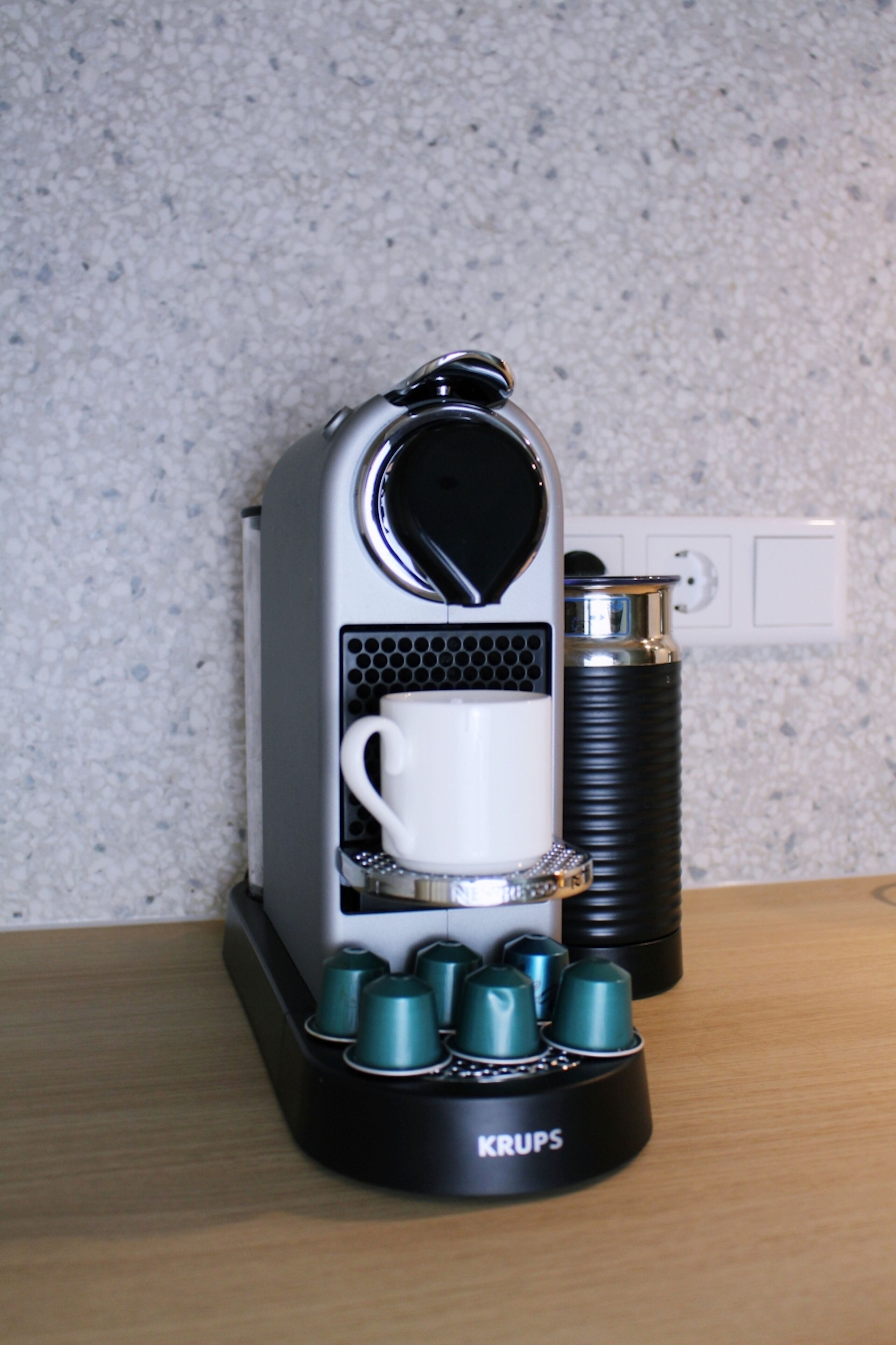 A Krups coffee machine with a white cup holder, a water reservoir, and a power outlet in the background.
