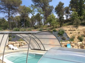 En swimmingpool med en glasvägg runt om, omgiven av en vackert anlagd trädgård med olika växter och en bergig sluttning i bakgrunden.