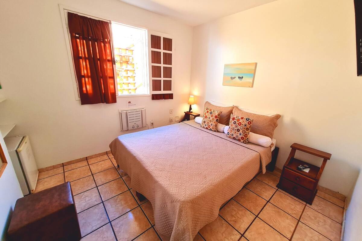 Une chambre à coucher avec un lit, une table de chevet, une lampe, une fenêtre avec des rideaux, un radiateur et un tableau au mur.