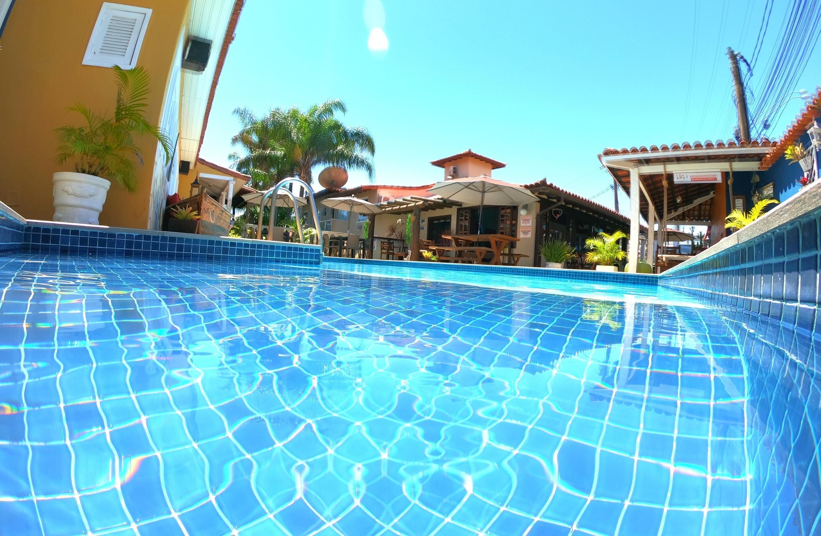 A swimming pool with tiled flooring, outdoor seating, and a covered area with umbrellas.
