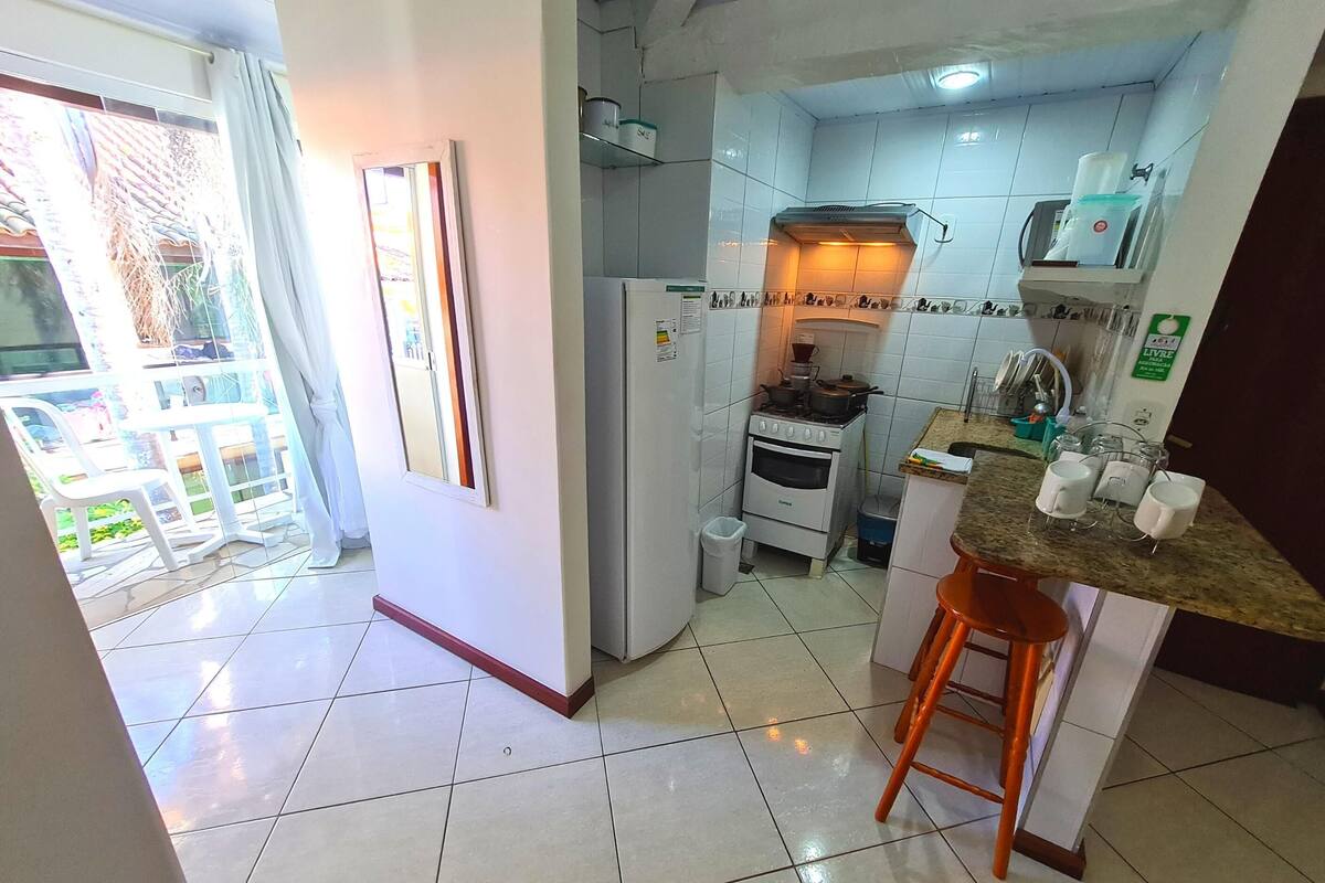 Une cuisine avec des meubles blancs, une cuisinière et un évier. Il y a une fenêtre avec un rideau, ainsi qu’un petit balcon avec une chaise et une table.