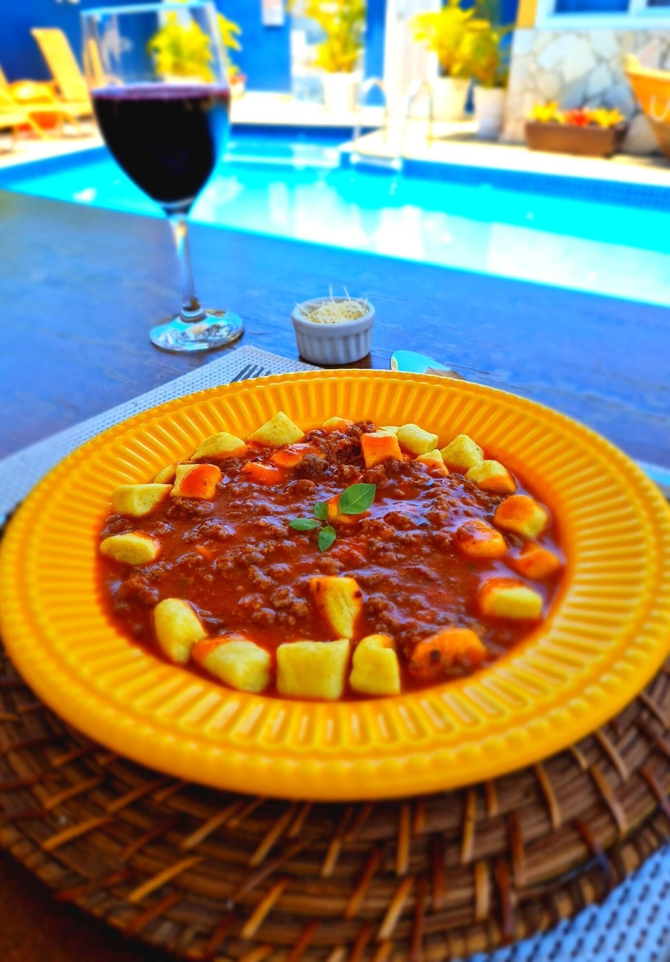A bowl of pasta with a red sauce and vegetables, a glass of red wine, and a poolside setting.