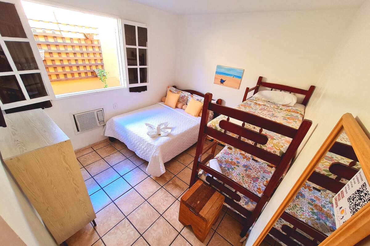 Une chambre avec un lit simple, un lit superposé en bois, une fenêtre avec des stores, un radiateur et une petite table.