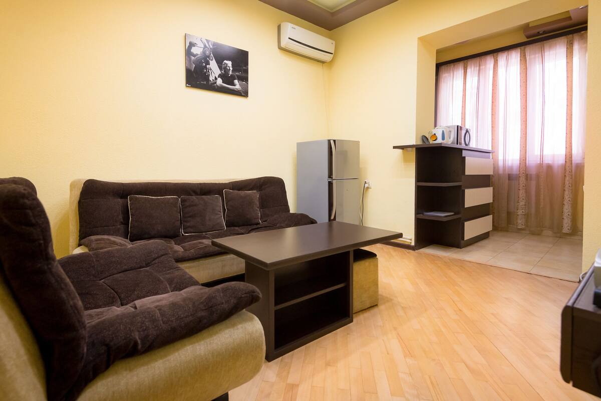 A compact living space with a sofa, a small table, a refrigerator, and a window with curtains.
