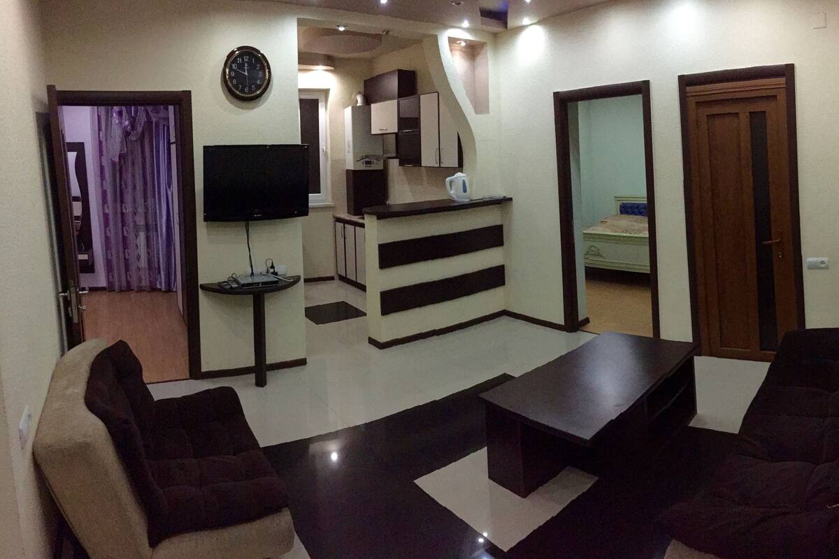 A living room with a sofa set, a coffee table, a TV, and a kitchen area in the background.