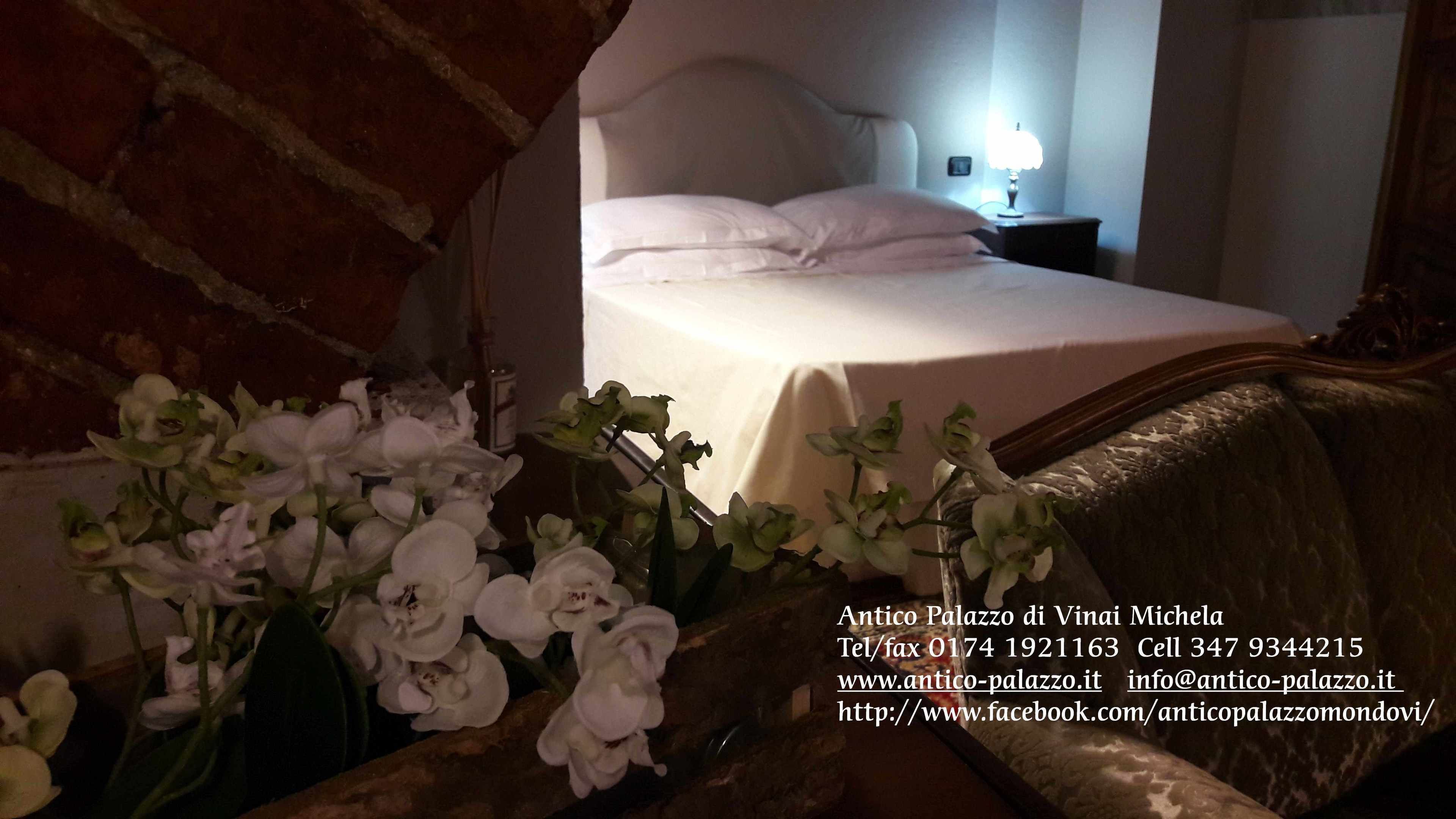 Een hotelkamer met een bed, witte kussens, een nachtkastje met een lamp en een vaas met witte bloemen.