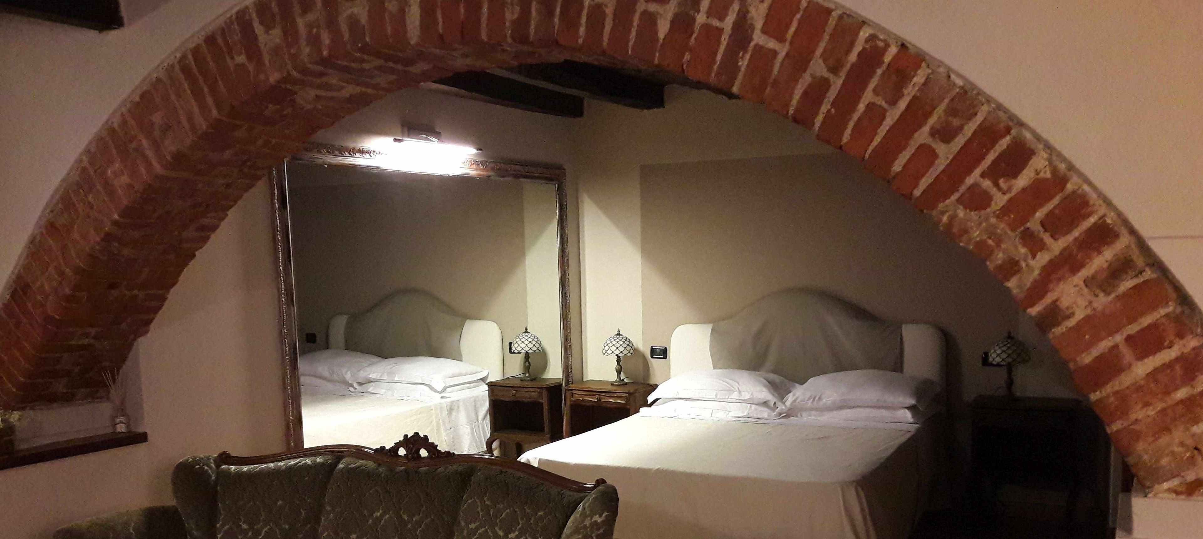 A room with two beds under a brick archway, a mirror, bedside tables with lamps, and a chair.