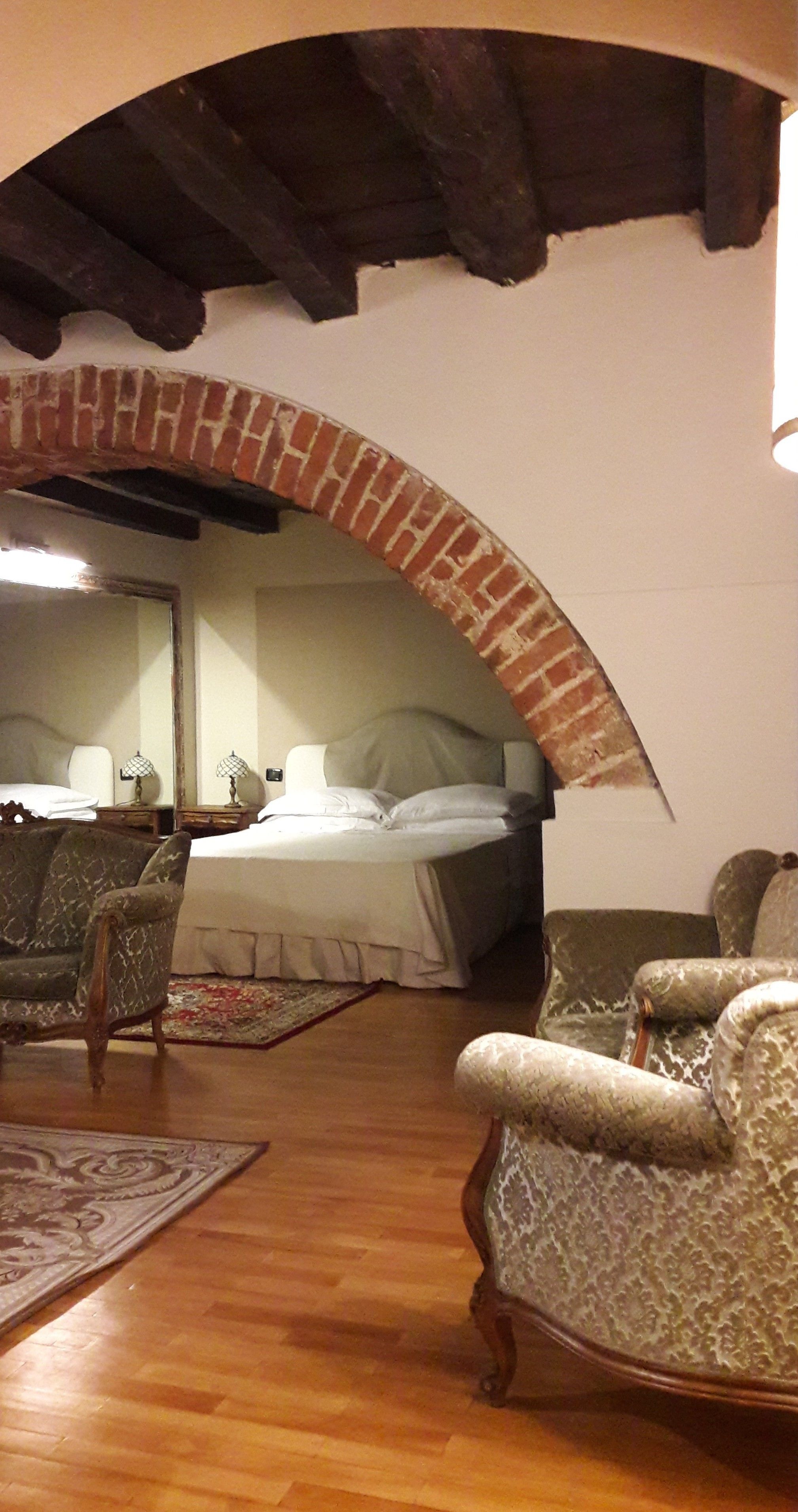 A room with a brick archway, wooden flooring, two upholstered armchairs, a bed with white linens, and a nightstand with a lamp.
