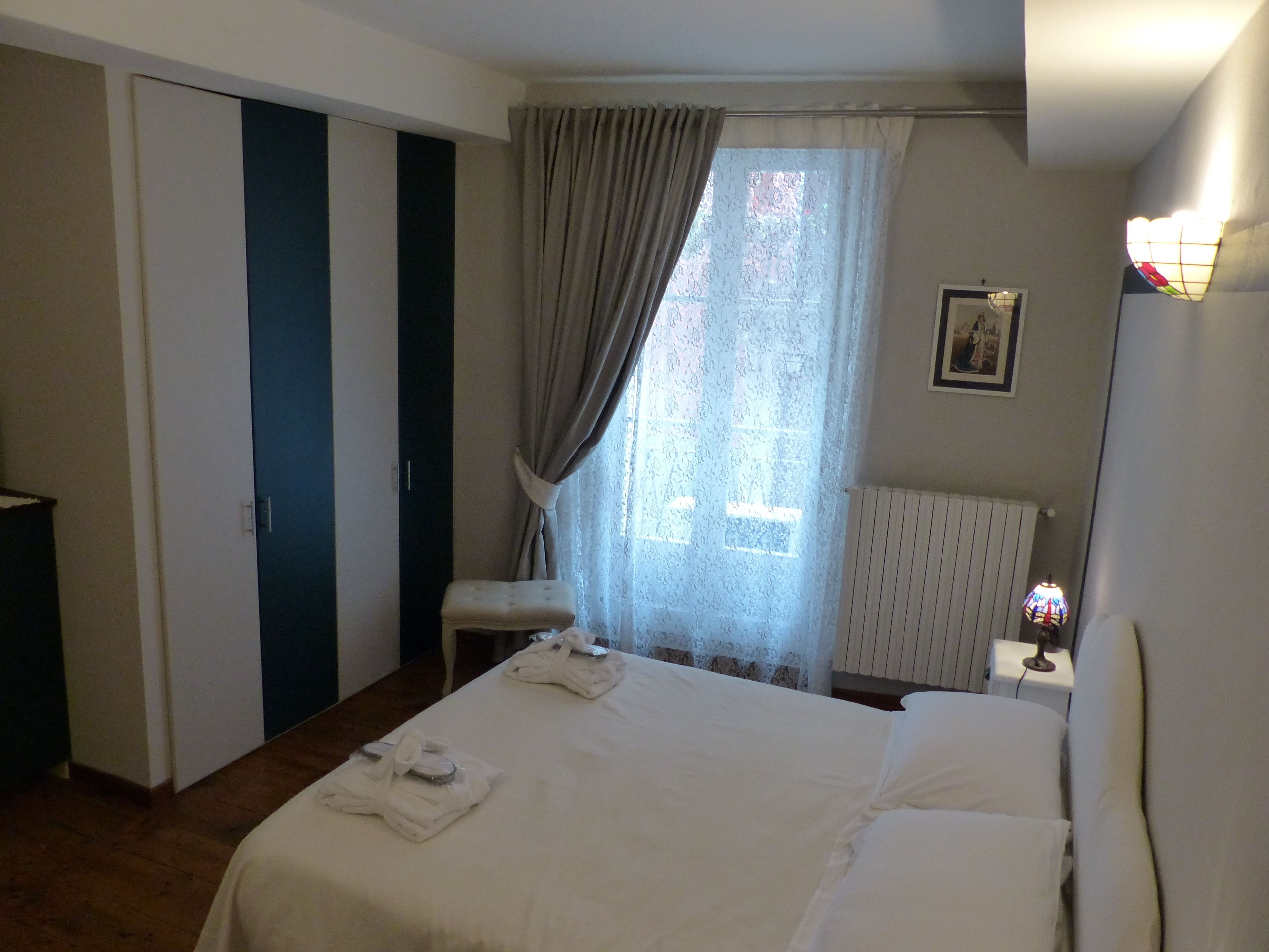 A hotel room with a bed, a nightstand, a lamp, a window with curtains, a radiator, and a door with a striped pattern.