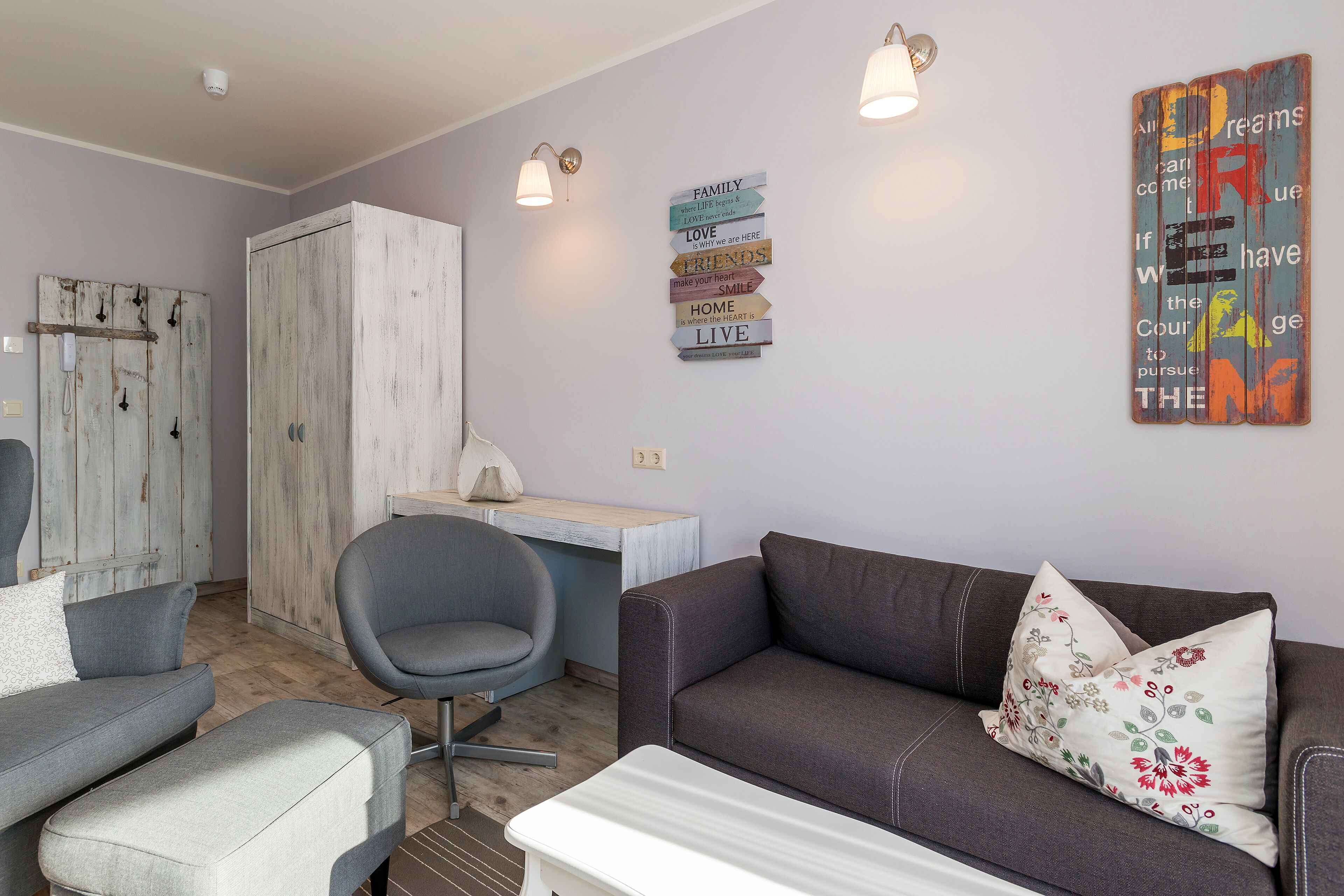 A living room with a grey sofa, a grey armchair, a wooden cabinet, and a small table. There are two wall-mounted light fixtures and a decorative sign with words like "family," "love," and "home."