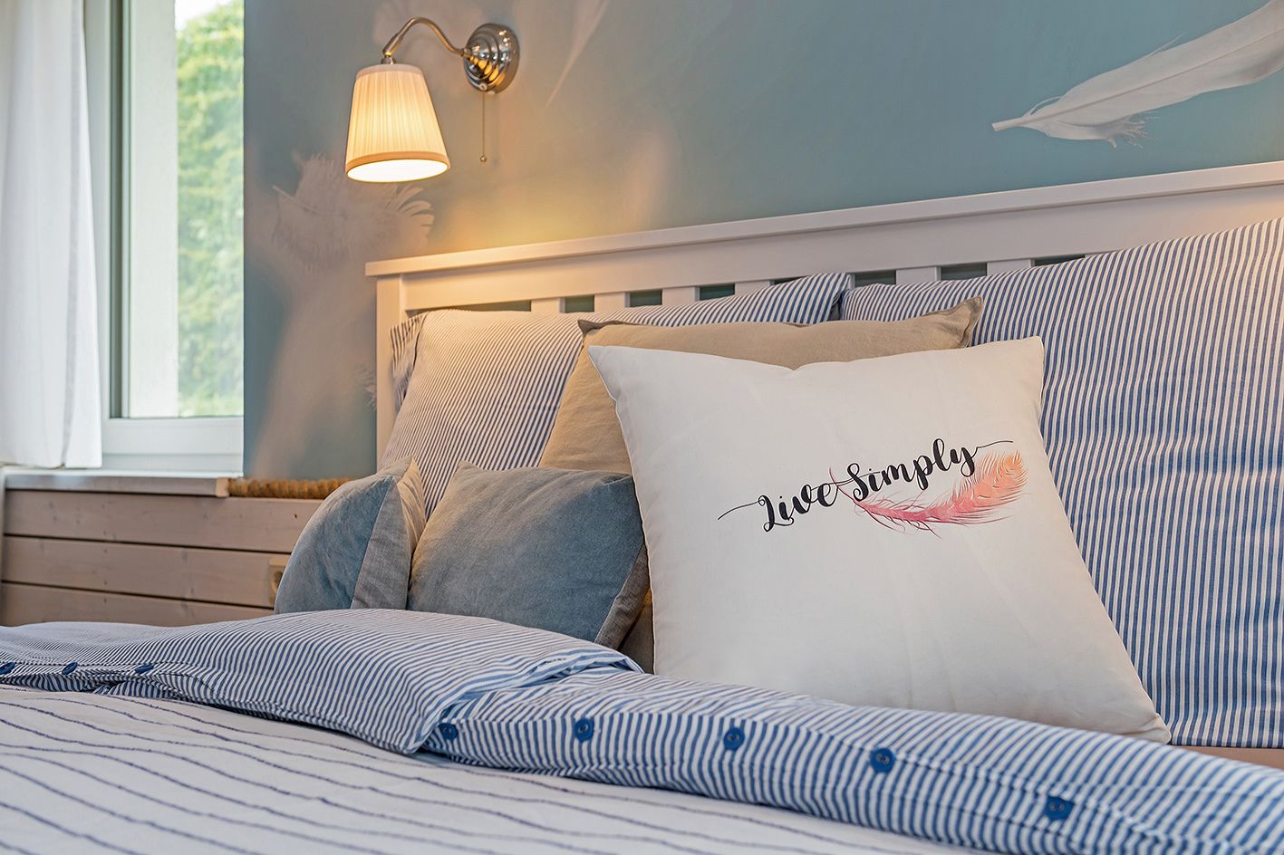 Ein ordentlich bezogenes Bett mit gestreifter Bettwäsche, ein Kopfkissen mit dem Aufdruck "Live Simply" und eine Wandlampe.