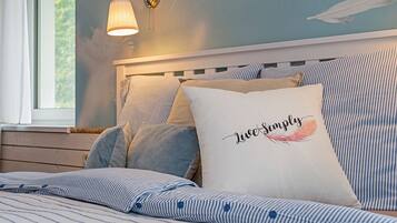 Ein ordentlich bezogenes Bett mit gestreifter Bettwäsche, ein Kopfkissen mit dem Aufdruck "Live Simply" und eine Wandlampe.