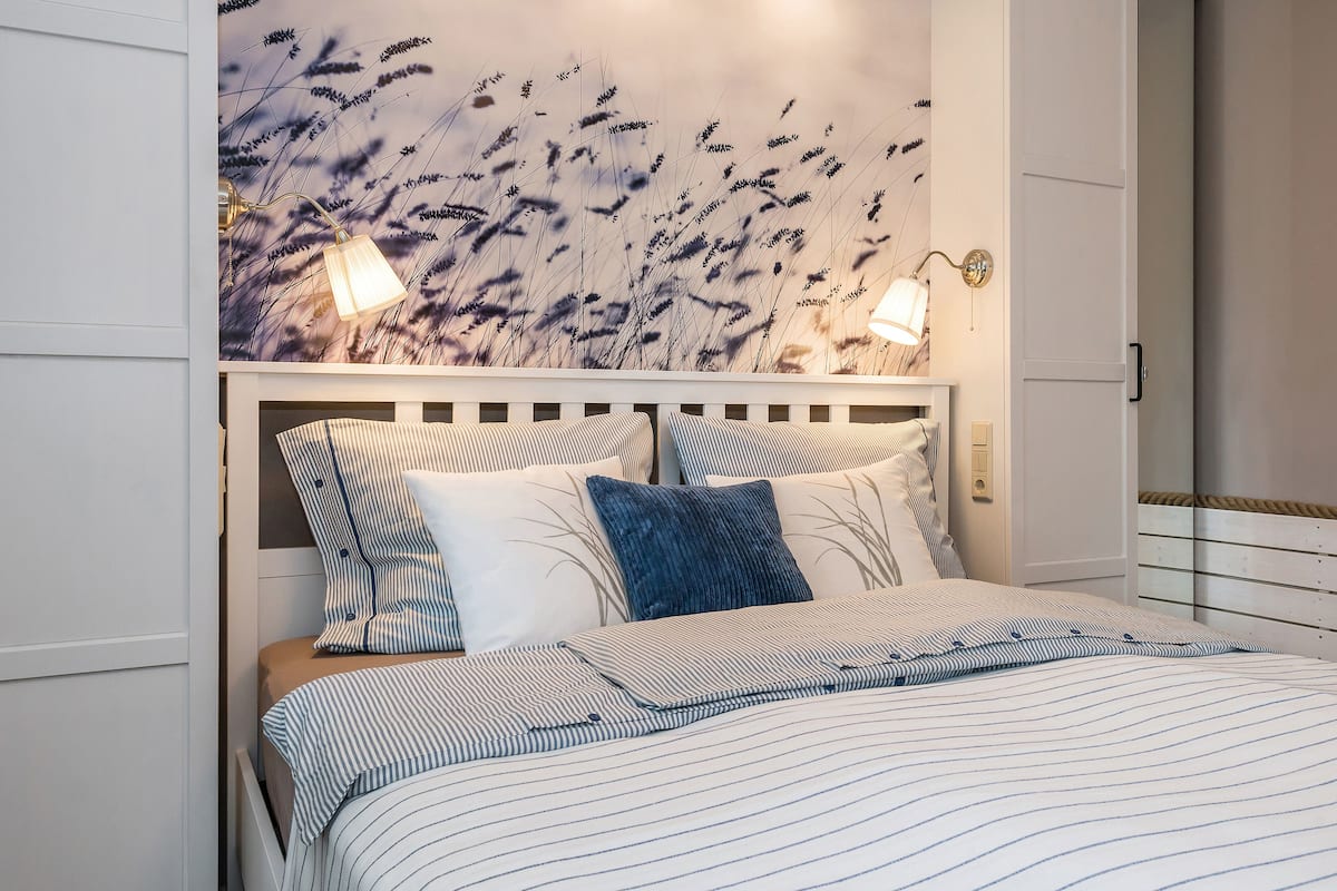 A neatly made bed with striped bedding, a wall with a floral mural, and two wall-mounted lamps.