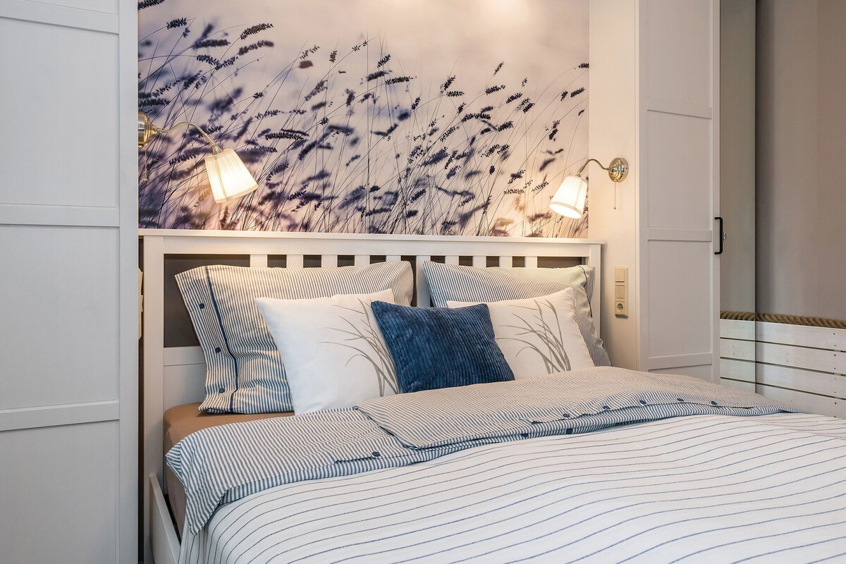 A bed with striped bedding, a headboard with a light fixture, and a wall with a floral mural.