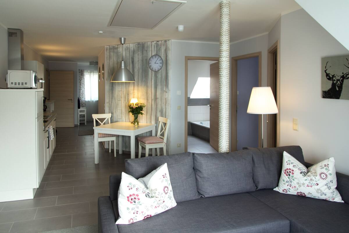 A modern living room with a grey sofa, floral cushions, a dining area with a table and chairs, and a kitchen in the background.