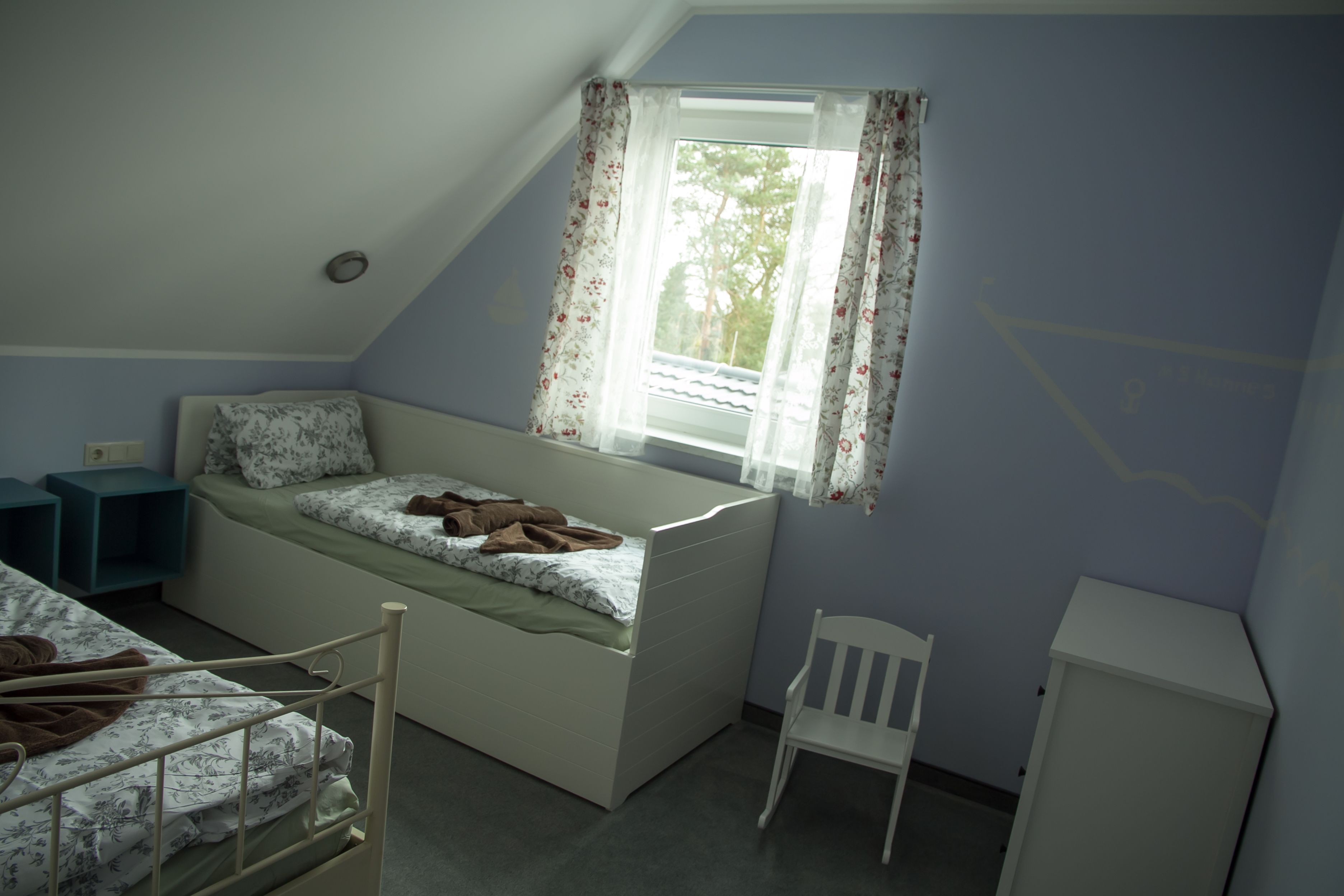 A bedroom with a bunk bed, a window with floral curtains, a chair, and a small cabinet.
