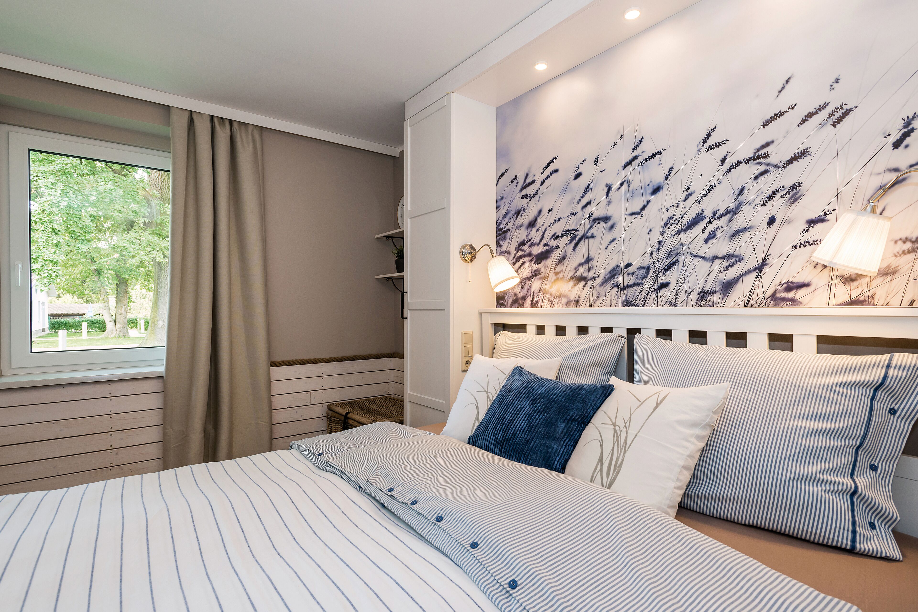 A bedroom with a large bed, a window with curtains, a wall-mounted lamp, and a wall art depicting tall grasses.