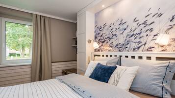 A bedroom with a large bed, a window with curtains, a wall-mounted lamp, and a wall art depicting tall grasses.