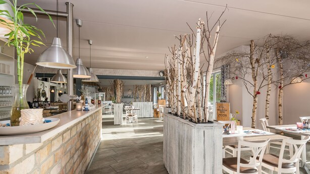 Ein geräumiges Restaurant mit einer langen Theke, hängenden Pendelleuchten und einem zentralen Pflanzgefäß mit Bäumen.