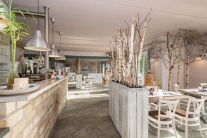 A spacious restaurant with a long counter, hanging pendant lights, and a central planter with trees. - Villa Meehr (Wusterhausen/Dosse)