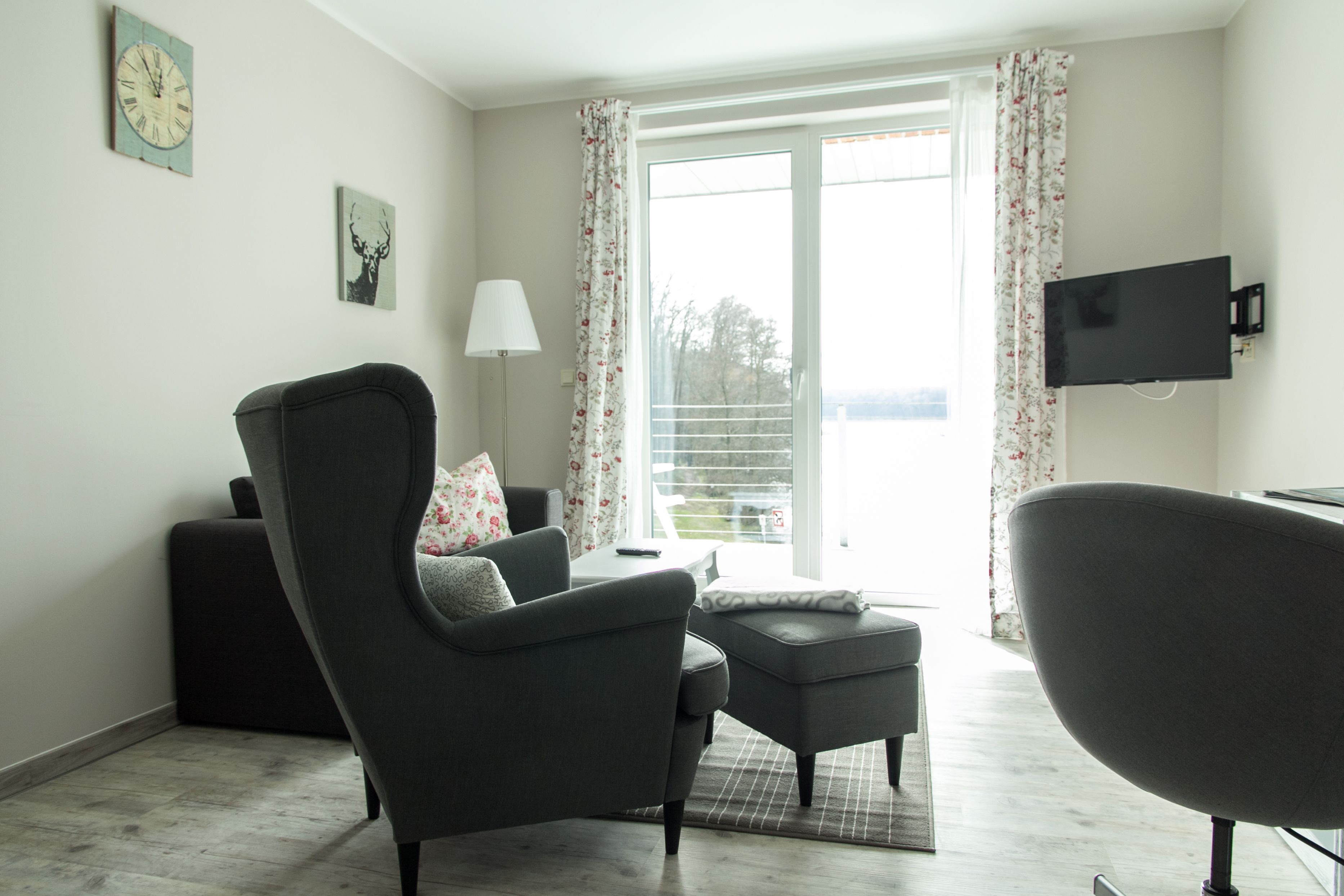 Ein modernes Wohnzimmer mit einer Couch, einem Sessel, einem Hocker, einem flachbildfernseher an der Wand und einem großen Fenster mit Vorhängen.
