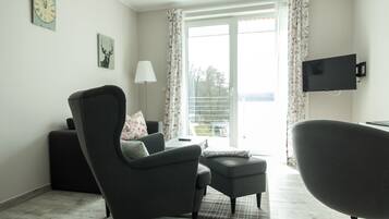 Ein modernes Wohnzimmer mit einer Couch, einem Sessel, einem Hocker, einem flachbildfernseher an der Wand und einem großen Fenster mit Vorhängen.