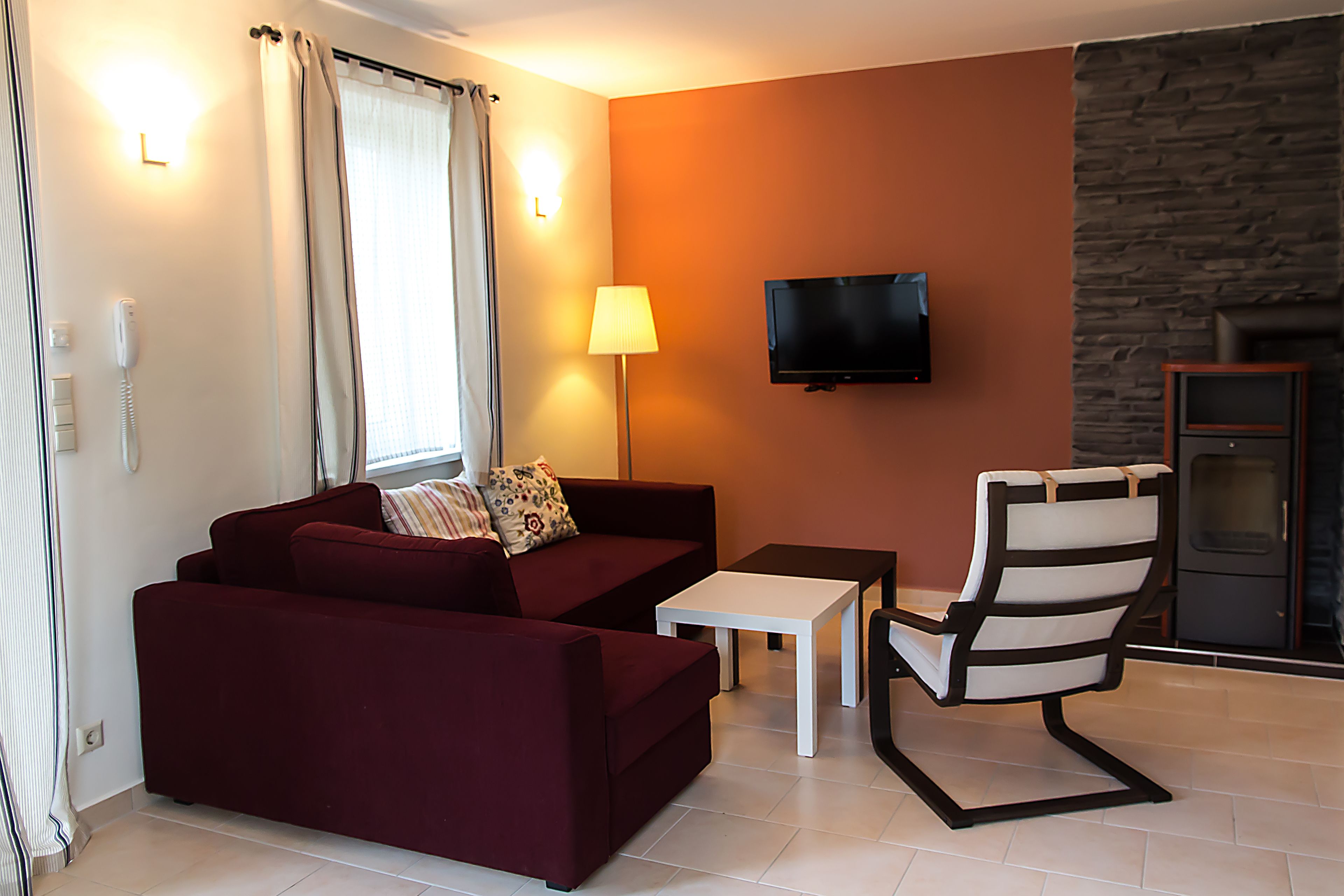 Una sala de estar con un sofá rojo, una mesa blanca, una silla a rayas, un televisor y una pared de piedra.