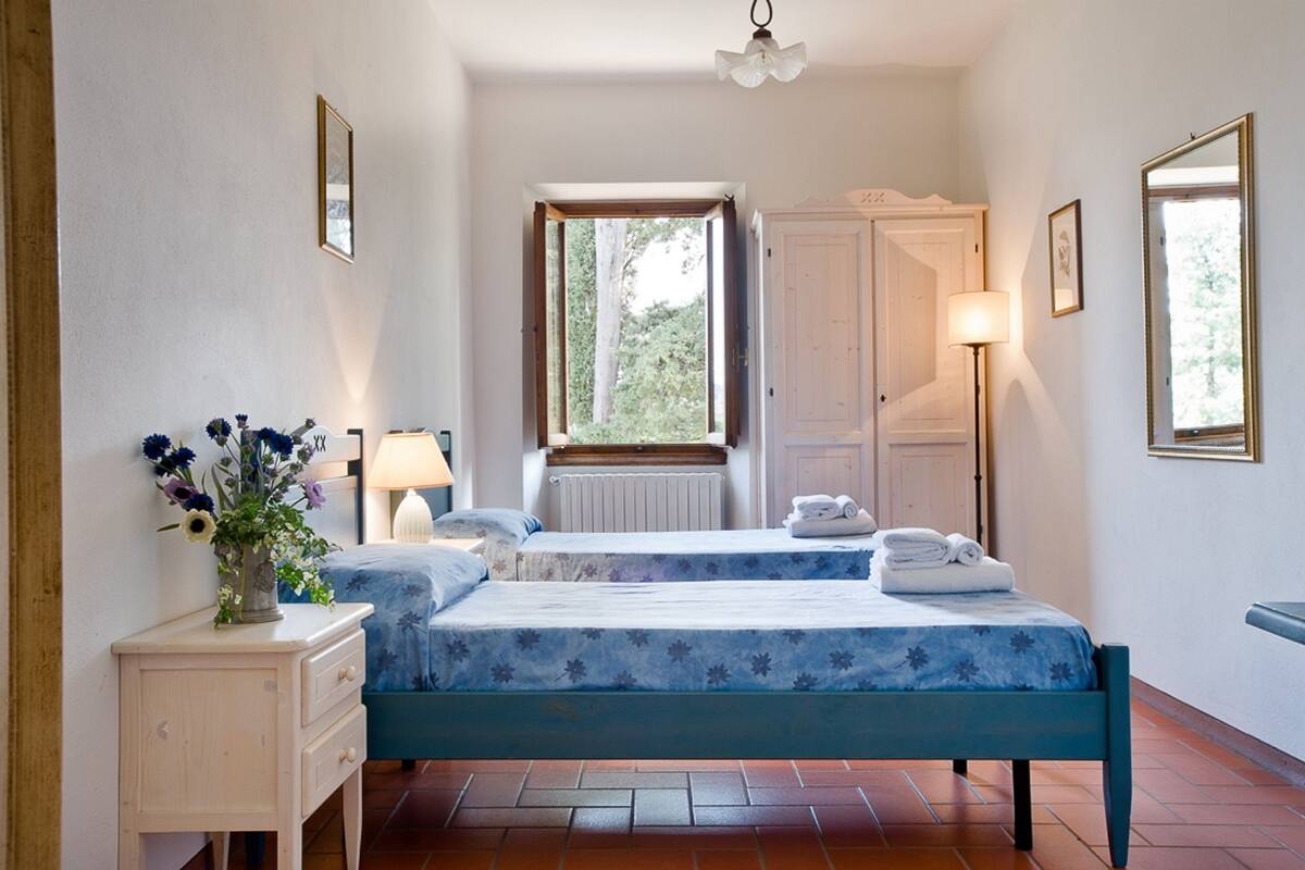A bedroom with two beds, a bedside table with a lamp, a window with curtains, a radiator, and a wardrobe.
